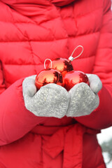 A woman in a red jacket and gray knitted mittens holds red Christmas balls.