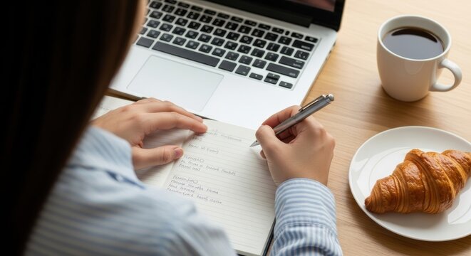 Female writing in notebook with coffee and croissant by laptop on wooden desk
