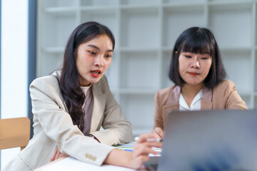 Two asian businesswomen working together, analyzing financial data on a laptop and discussing strategy in a modern office
