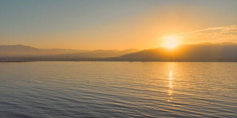 Obraz premium Peaceful sunrise over calm waters with a warm golden glow reflecting on the gentle waves and distant mountains in the background.