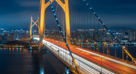 Obraz premium Vibrant night view of a large cable-stayed bridge with light trails from cars and a glowing city skyline in the background.