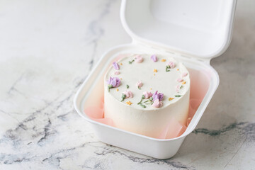 Delicate small bento cake with white cream cheese frosting in a gift box. Trendy cake as a gift for a beloved one. Spring tender cake for a woman decorated with flowers on the white background.