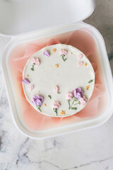 Delicate small bento cake with white cream cheese frosting in a gift box. Trendy cake as a gift for a beloved one. Spring tender cake for a woman decorated with flowers on the white background.
