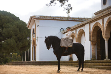 Young horsewoman riding a black Spanish thoroughbred horse next to an old hermitage. She has a...