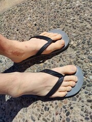 Bare feet in worn black flip-flops on a sunlit pavement, symbolizing a carefree summer stroll. Metaphorically, the simple footwear reflects a journey of ease, grounded freedom, and the shedding.