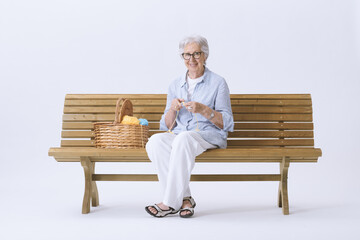 A smiling and happy elderly woman sitting on a bench is knitting and looking at the camera.