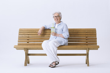 Fototapeta premium Smiling and happy elderly woman sitting on a bench holding a glass of water, looking at the camera.
