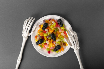 Skeleton hands holding plate of colorful jelly beans and black small decorative skulls on a gray background