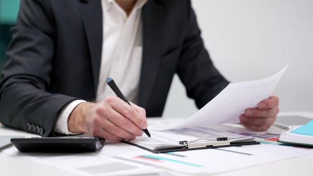 Close up of a male hand filling out documents with a pen using a calculator sitting at a desk at a workplace in a business office. Businessman is engaged in paperwork, making financial calculations
