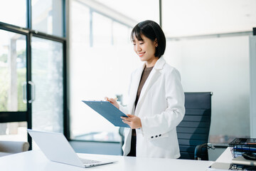 Asian businesswoman in blazer reviewing report on clipboard while standing at desk. Confident,...
