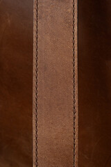 A high-quality, close-up shot of a textured brown leather strap with visible stitching, running vertically against a dark brown leather background