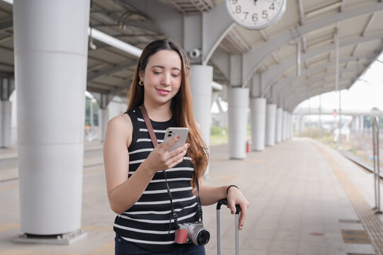 A smiling young woman is standing on a sky train platform, using her smartphone while waiting for the train.