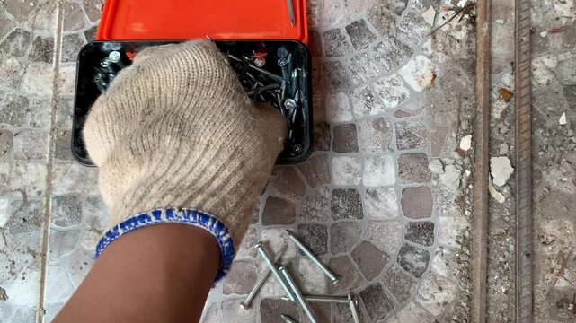 4K close-up of a gloved hand taking shiny steel nails from a black box onto tiled floor, sharp focus on tools. Great for repair, DIY, construction, hardware, and industry themes.