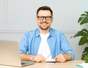 A smiling, bespectacled man in a blue shirt sits at a desk with a laptop, looking directly at the camera, radiating friendliness in a bright office setting