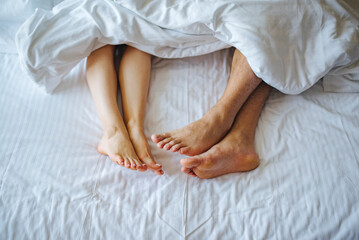 Close-up of a couple’s bare feet under a white blanket on a bed. Relaxed and intimate atmosphere symbolising love, comfort