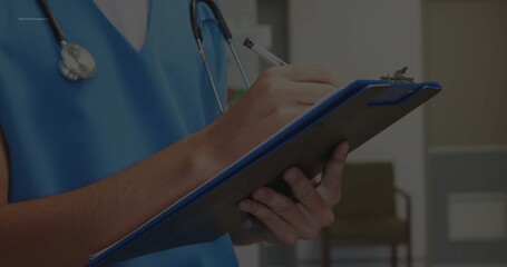 Clinician holding clipboard and writing notes in clinic hallway with scrubs and stethoscope