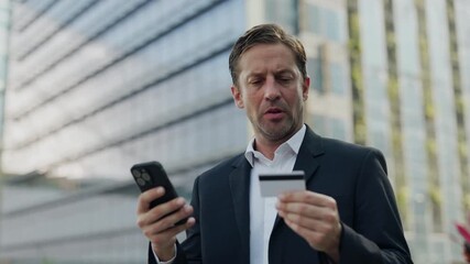 Portrait of focused entrepreneur transfers money with smartphone and credit card on city street. Successful businessman uses banking app for payment standing near office building - Powered by Adobe