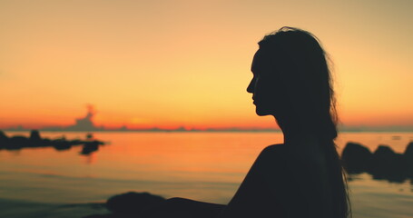 A silhouette of a woman sitting on a sandy beach, framed against the vibrant hues orange sunset in Thailand. The calm waters reflect the colorful sky as she gazes out towards the horizon.