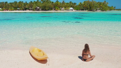 Aerial view of a young woman sitting on the sand of a pristine tropical beach, with a yellow kayak next to her and crystal clear turquoise water in the background