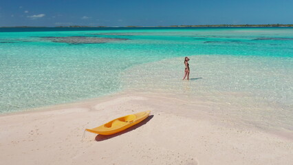 Woman enjoying a tropical beach vacation, walking on a sandbar with a kayak nearby, surrounded by turquoise ocean and blue sky on a sunny day