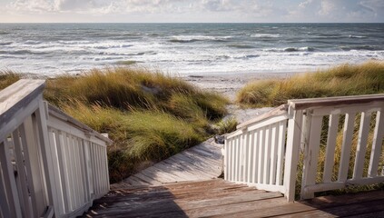 Coastal walkway down to a sandy beach. Ocean waves cresting in the distance. Wooden stairs and white railing lead to the shore. Coastal dunes and tall grasses