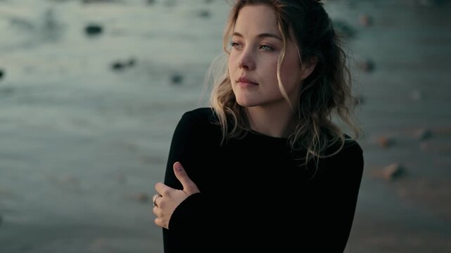 A thoughtful woman stands on a chilly beach, embracing herself to keep warm on a cold day. The serene landscape adds to the contemplative mood.