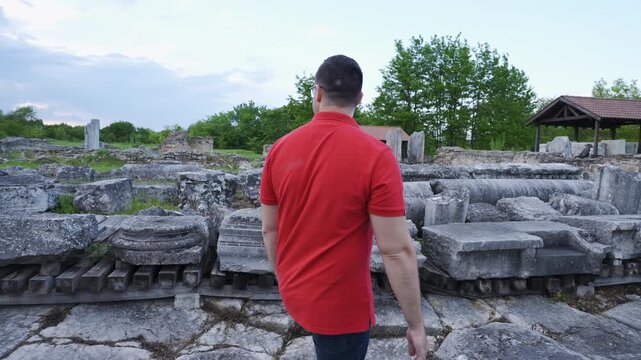 Follow shot of a man in red shirt walking through the Roman ruins of Nicopolis ad Istrum during sunset with clouds in the distance