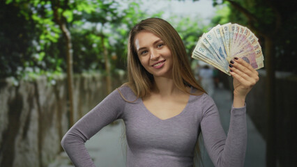 Young woman holding polish zloty banknotes outdoors on a city street, smiling confidently at the...