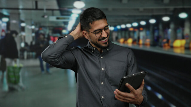 Young man using a tablet in a busy indoor train station, smiling, wearing glasses, with people in the background, illustrating modern travel and communication.