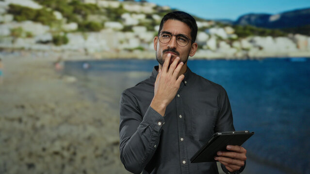 Young man with a thoughtful expression holding a tablet on a beach background with the sea in view.