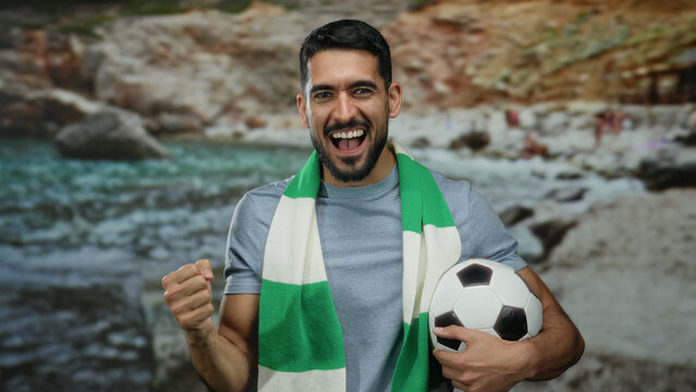 Young man at seaside beach holding soccer ball wearing green scarf looking enthusiastic and cheerful.