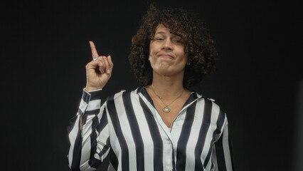 Woman wearing black and white striped blouse and gold pendant necklace pointing finger upward in...