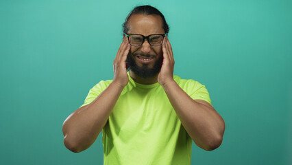 Man pressing temples with both hands while wearing glasses and neon green shirt in studio teal backdrop; headache pain.