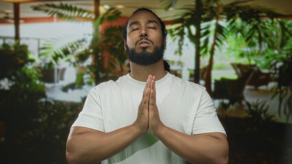 Man praying with hands pressed together in hotel building lobby amid indoor plants and seating;...