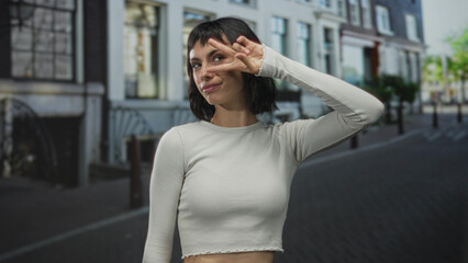 Obraz premium Woman shows v sign with hand to face while smiling on a sunlit cobblestone street; carefree happiness.