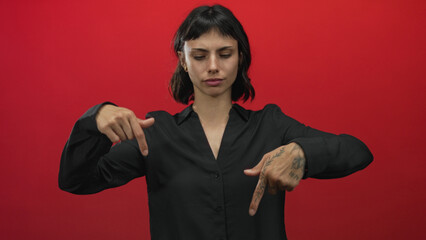 Young hispanic woman pointing finger down with stern expression and tattooed hand in red studio...