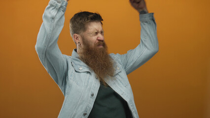 Man with beard and open mouth wearing denim jacket raises clenched fists in orange studio;...