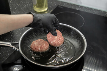 A gloved hand meticulously places raw meat cutlets into a hot frying pan on a modern induction hob,...