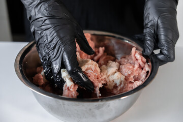 Chef hands in black protective gloves thoroughly mixing raw minced pork and beef in a stainless...