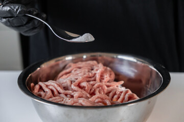 Chef's gloved hand carefully sprinkles essential salt seasoning onto fresh raw minced meat in a sleek metal bowl, preparing delicious homemade cutlets or meatballs