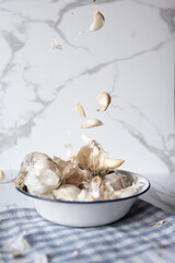 Garlic cloves and bulbs with roots falling into a white enamel bowl on a blue and white checkered tablecloth, marble table background
