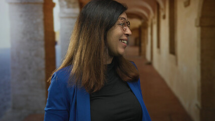 Woman wearing blue jacket and glasses smiles while turning head in sunlit building corridor;...