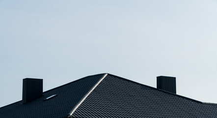 Modern house roof with dark grey tiles, two black chimneys, and a skylight against clear blue sky