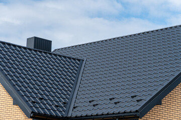 Fototapeta premium Modern dark metal tile roof with chimney and light brick wall under a blue sky.