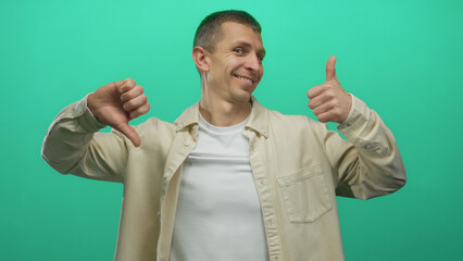 Man wearing beige shirt and white tee stands against green background displaying conflicting thumbs...