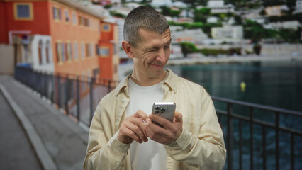 Man using smartphone outdoors on a seaside promenade with a scenic view of the sea and buildings.