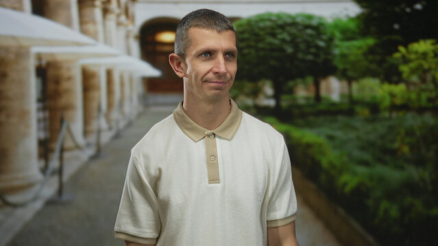 Caucasian man smiling outdoors on a sunny day in a landscaped urban street setting with lush greenery and classical architecture in the background.