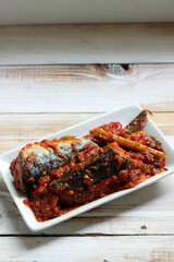 Sarden or Sardencis. Spicy Indonesian mackerel dish served in rich red chili sauce with lemongrass on a white plate over rustic wooden table.