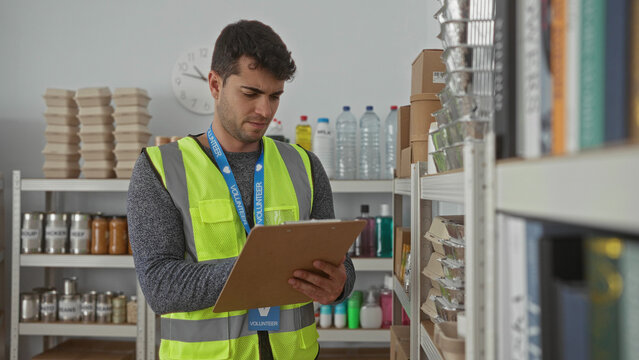 Man holds clipboard while writing inventory in warehouse as male young hispanic volunteer organizes donation food.