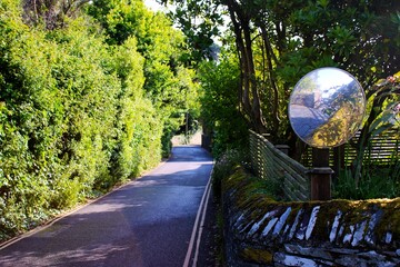 Country lane bordered by green hedges, with a convex traffic mirror mounted on a stone wall.